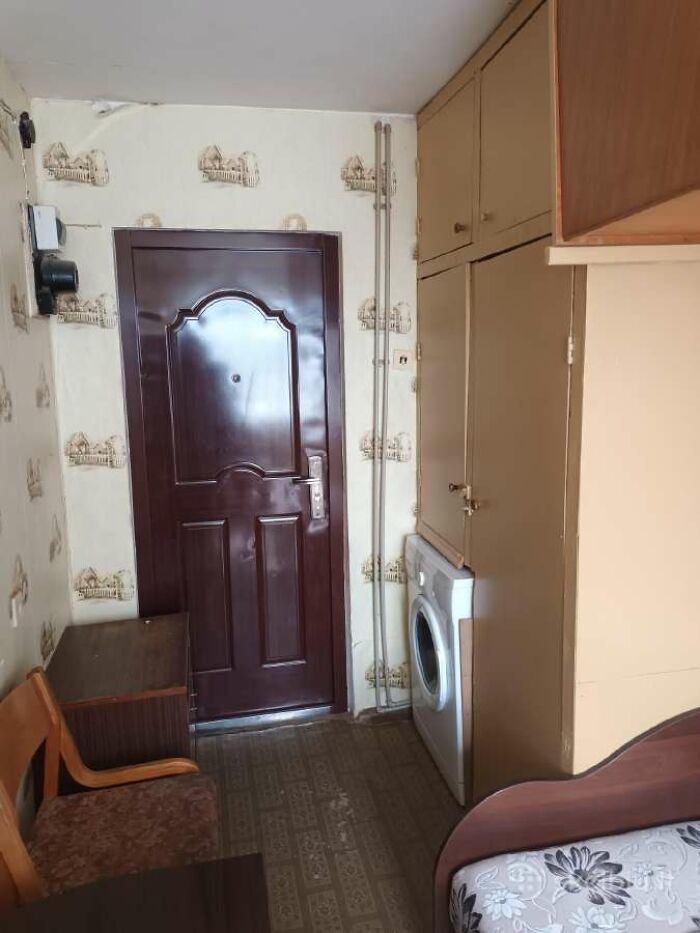 Small cluttered room with dark door, built-in cabinets, washing machine, and mismatched furniture in real estate listing.