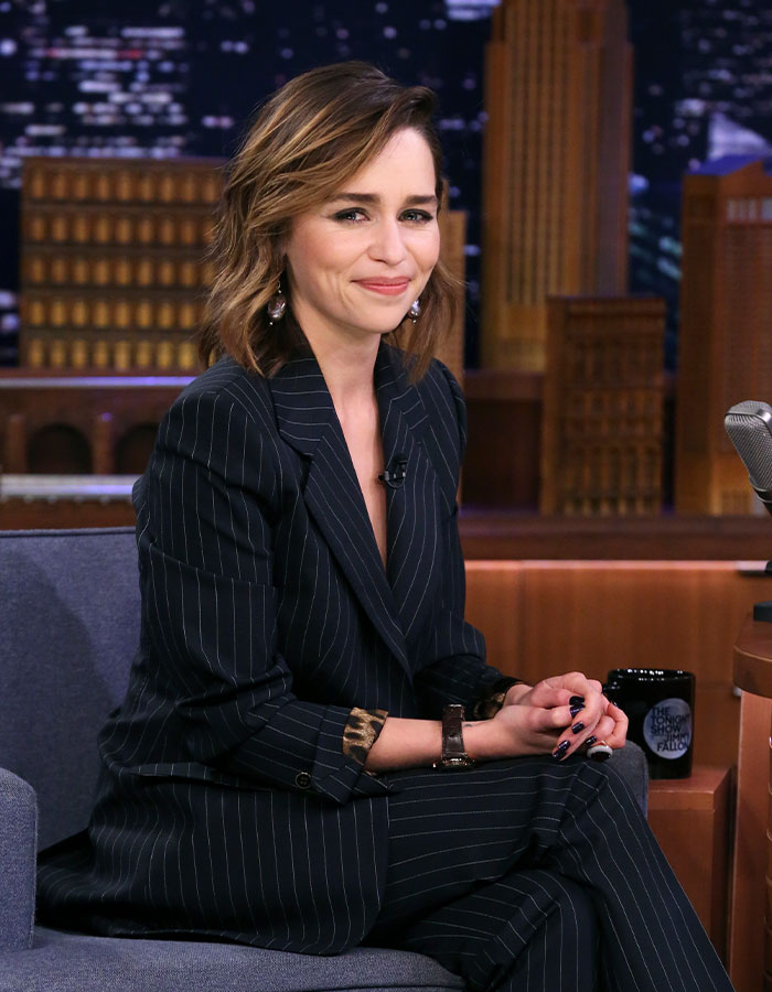 Emilia Clarke smiling during a talk show interview, wearing a pinstripe suit, highlighting Hollywood face and surgeries discussion.