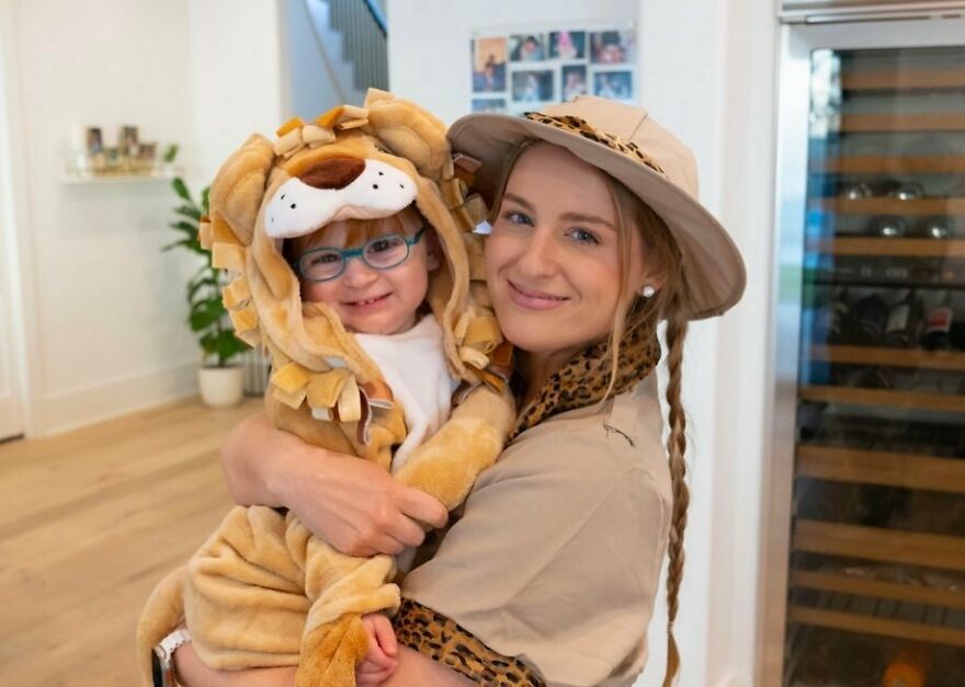 Meghan Trainor smiling and holding a child dressed in a lion costume in a bright, modern home setting.