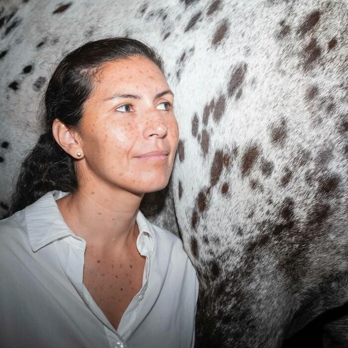 Woman with freckles and dark hair closely posing next to a spotted horse, showing pets and their humans looking alike.