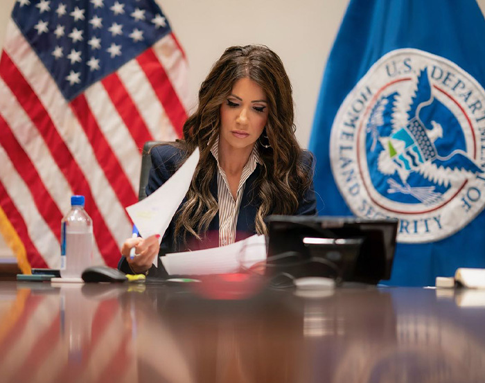 Kristi Noem working at a desk with American flag and Homeland Security emblem in the background, showing transformation. Kristi Noem working at a desk with American flag and Homeland Security emblem in the background, showing transformation.