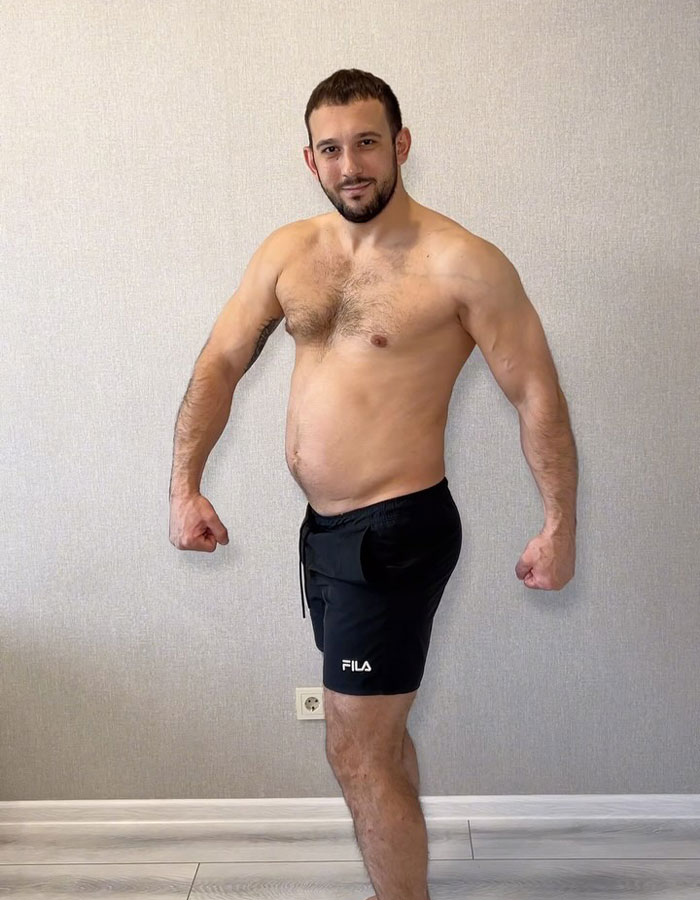 Shirtless fitness coach posing indoors wearing black shorts, showing muscular arms and slight belly against plain wall background.