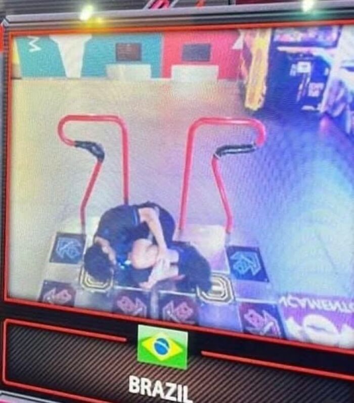 Two people bending over on a dance arcade machine in an indoor gaming area with Brazil flag visible.