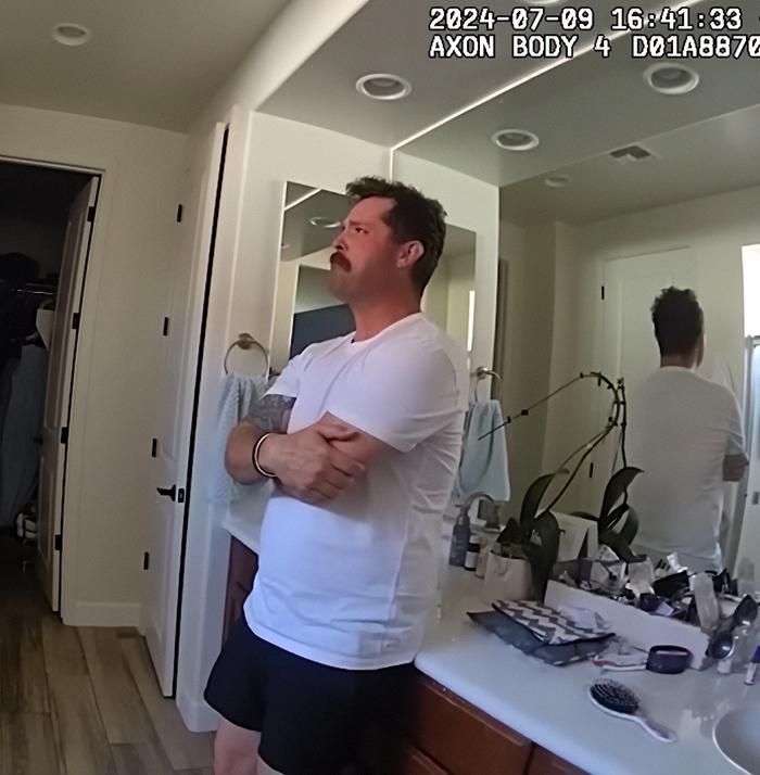 Man in white shirt and black shorts standing in a bathroom with arms crossed, related to dad leaving 2YO in car incident. Man in white shirt and black shorts standing in a bathroom with arms crossed, related to dad leaving 2YO in car incident.