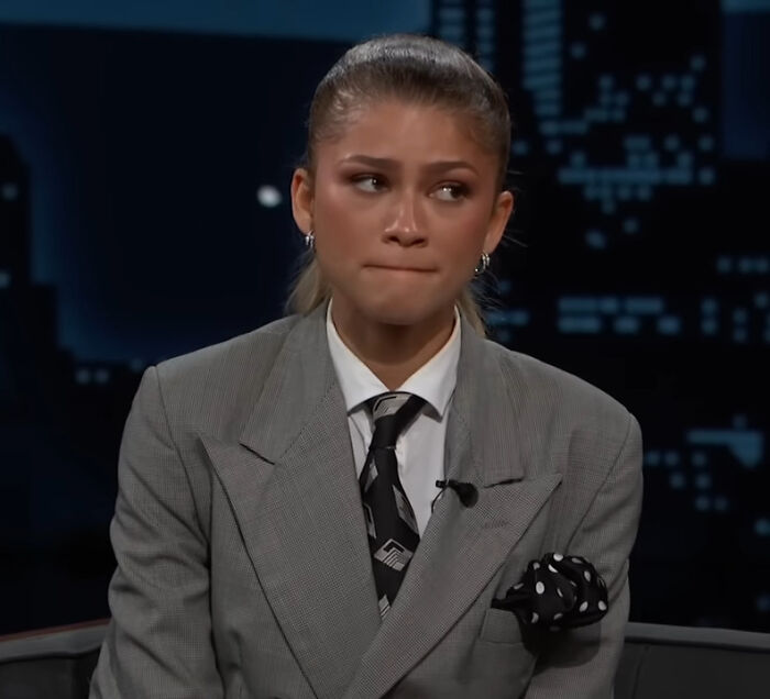 Zendaya in a gray suit and patterned tie, appearing thoughtful during a late night TV interview about petty feud controversies.