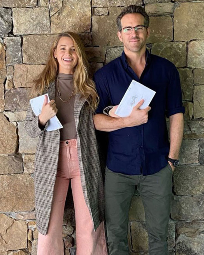 Justin Baldoni and Blake Lively standing together holding papers against a stone wall background.