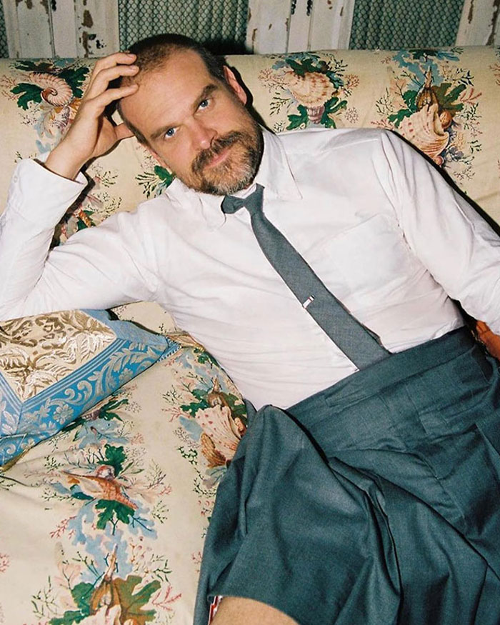 Man with a beard reclining on floral couch in white shirt and gray tie, contrasting Lily Allen&rsquo;s CFDA fashion look debate online.