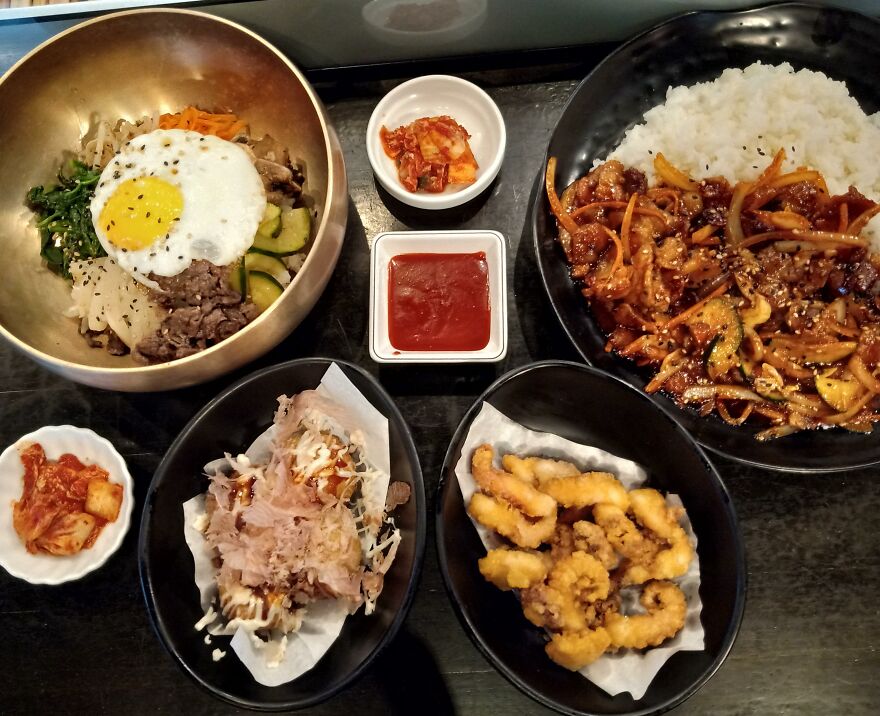 Asian cuisine spread including bibimbap with egg, stir-fried dish with rice, fried calamari, and kimchi side dishes on a table