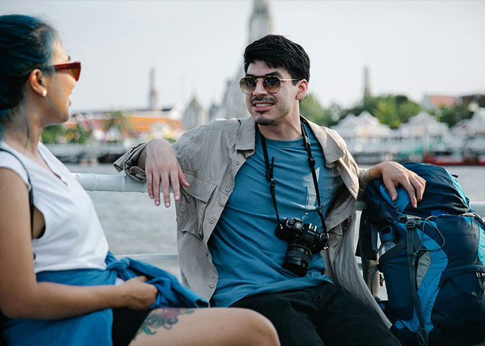 Two travelers wearing sunglasses relax and chat by the waterfront with a camera and backpack nearby in a casual setting.