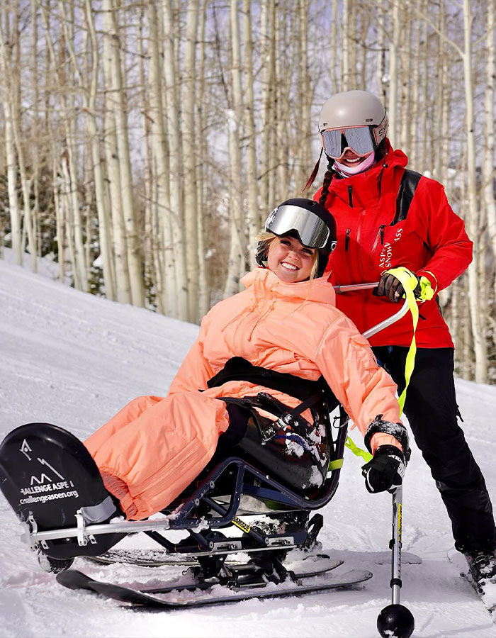 Girl who was paralyzed at 16 enjoying adaptive skiing with an instructor on a snowy slope surrounded by bare trees.
