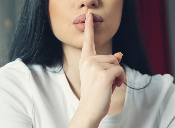 Close-up of a woman with long dark hair holding her finger to her lips in a gesture of girl code secrecy.