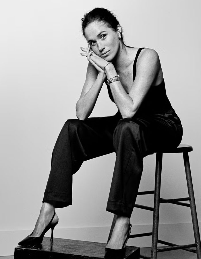 Meghan Markle sitting on a stool in black outfit and heels, posing thoughtfully in a black and white editorial photo.