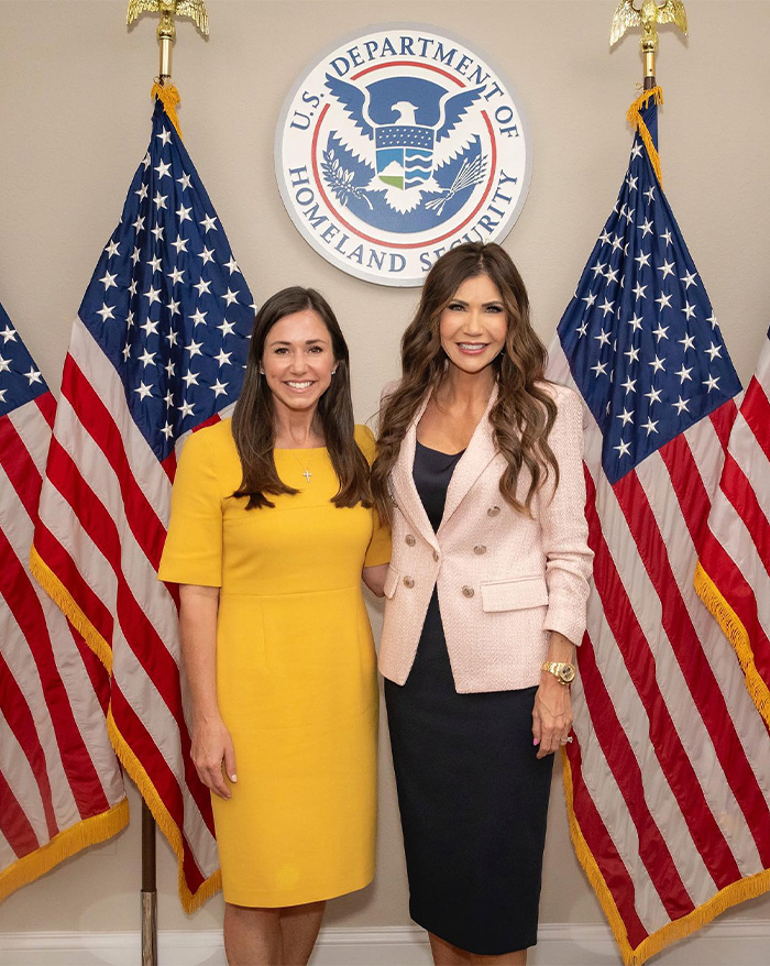 Two women posing in front of U.S. Department of Homeland Security sign with American flags, showcasing Kristi Noem transformation. Two women posing in front of U.S. Department of Homeland Security sign with American flags, showcasing Kristi Noem transformation.