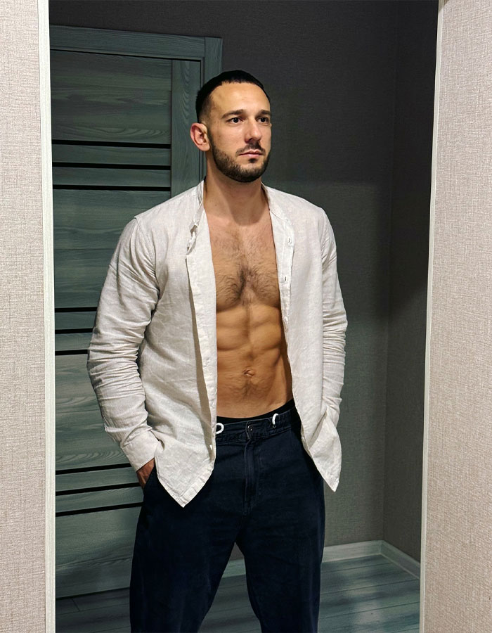 Fitness coach with visible abs wearing an open white shirt and dark pants standing indoors against a gray wall.