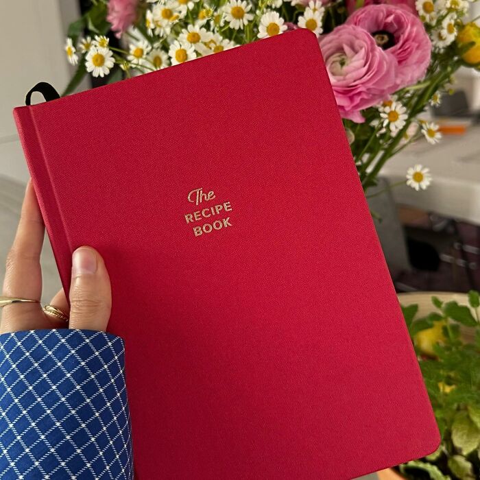 Hand holding a red recipe book with flowers in the background, perfect gifts for an aesthetic personality.