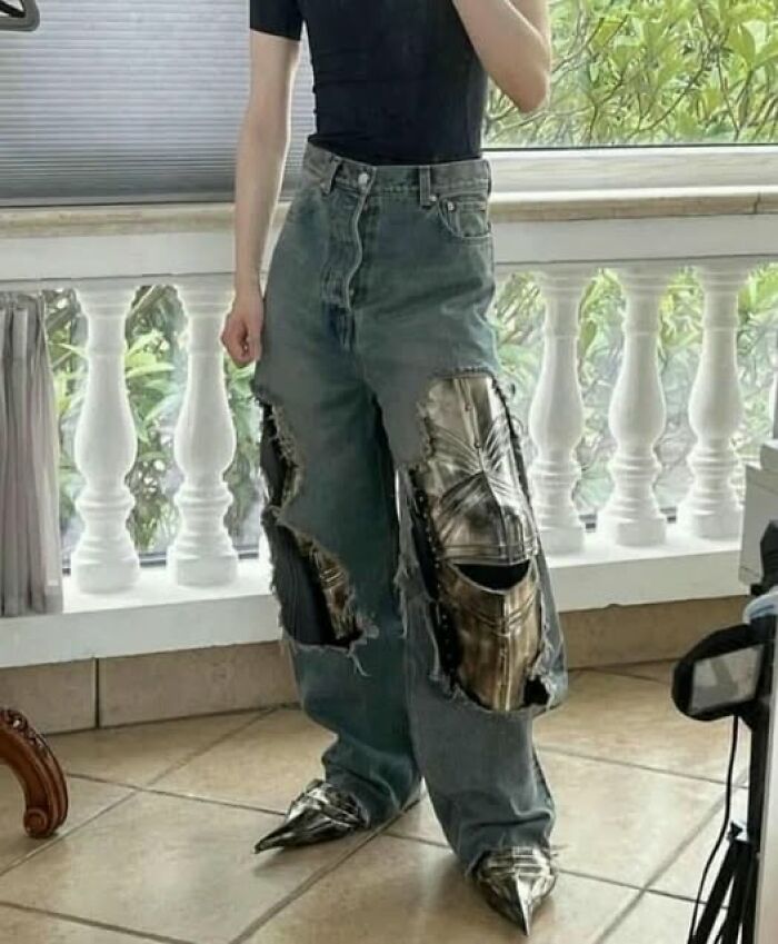 Person wearing pics that go hard ripped jeans with armor patches and pointed metal shoes standing indoors near balcony railing.