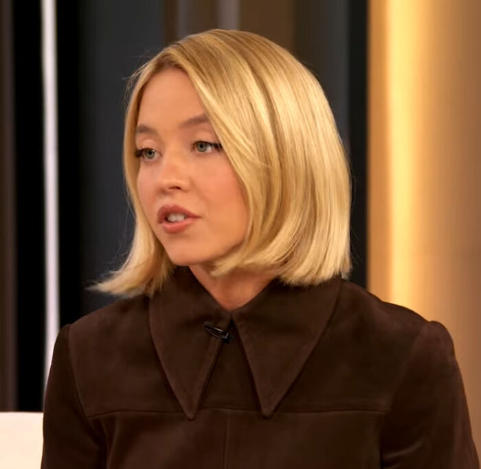 Sydney Sweeney speaking in an interview, wearing a dark brown top with a large collar, discussing latest controversy.