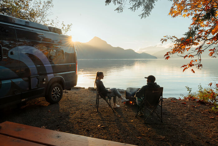 Two people sitting by a campfire near a van, reflecting on their journey and purpose beside a calm lake at sunset.