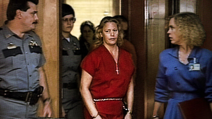 Woman in red prison uniform escorted by police officers in a courtroom scene from a Netflix doc about a famous serial criminal. Woman in red prison uniform escorted by police officers in a courtroom scene from a Netflix doc about a famous serial criminal.