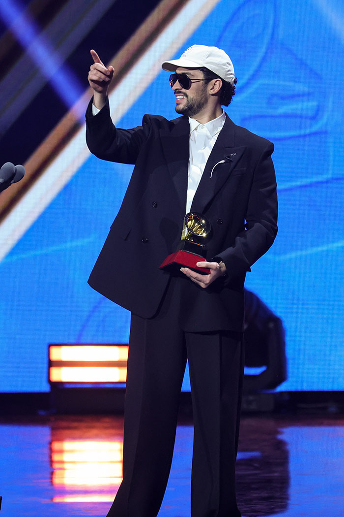 Male artist at 2025 Latin Grammy Awards wearing oversized suit and white cap, holding a trophy on stage, worst dressed stars.