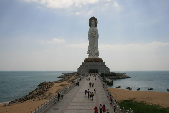 Massive coastal sculpture of a standing figure with visitors walking along a long pier under a cloudy sky.