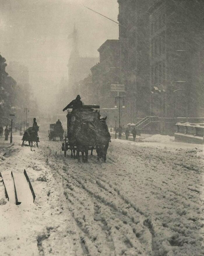 Horse-drawn carriages traveling through a snowy Victorian-era city street during heavy snowfall.