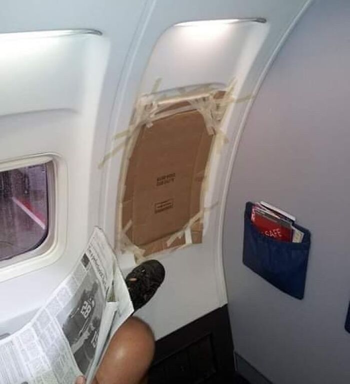 Passenger reading a newspaper with a taped cardboard sign over airplane emergency exit in an aviation meme setting.