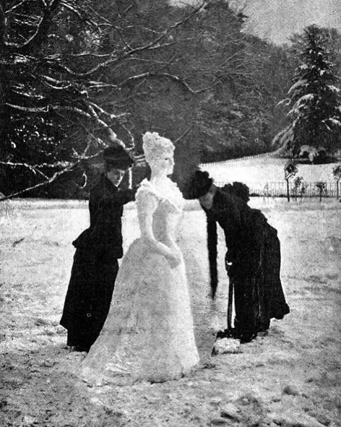 Two Victorian women building a snow sculpture of a woman in a white dress in a snowy outdoor setting, weird Victorian era photo.