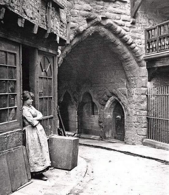 Victorian era street scene with a woman standing outside a wooden door near stone archways and historic buildings.