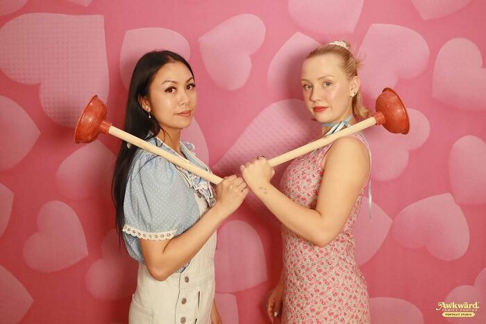 Two women posing with plungers on a pink heart background in an awkward studio pic showing playful cringe moments.