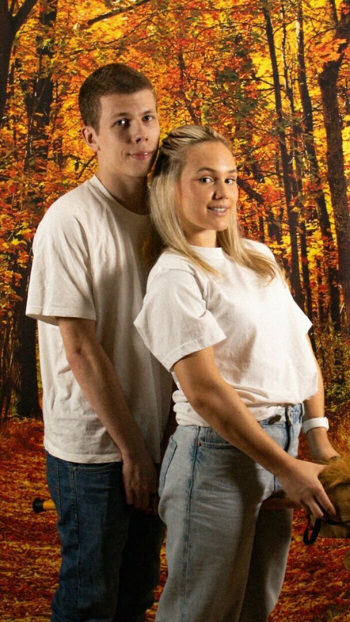 Young couple posing awkwardly in a studio with an autumn forest backdrop, wearing casual white shirts and jeans.