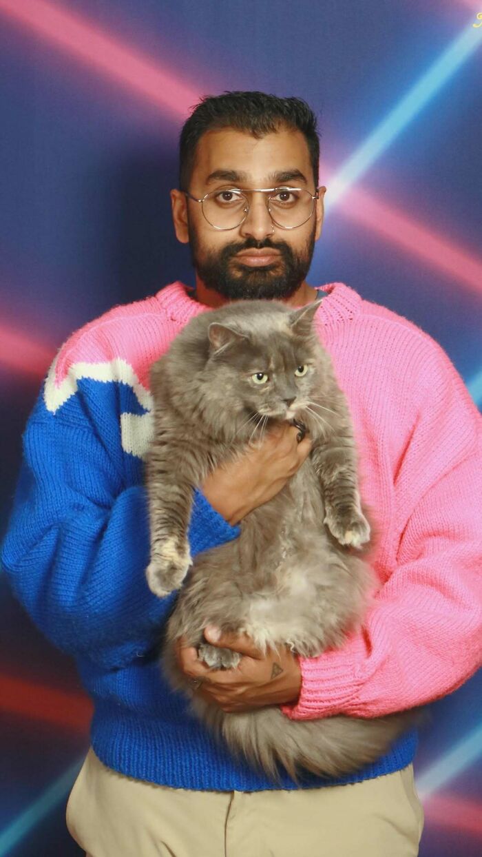 Man in colorful sweater holding large gray cat in front of a retro laser light backdrop in an awkward studio pic.