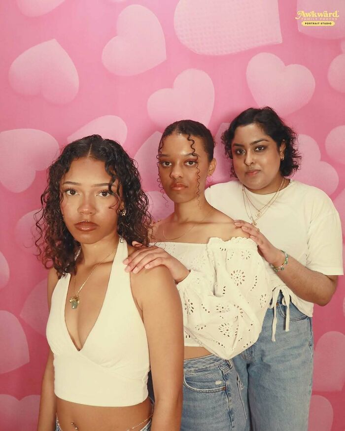 Three young women posing awkwardly in front of a pink heart-patterned backdrop in an awkward studio photo setting.