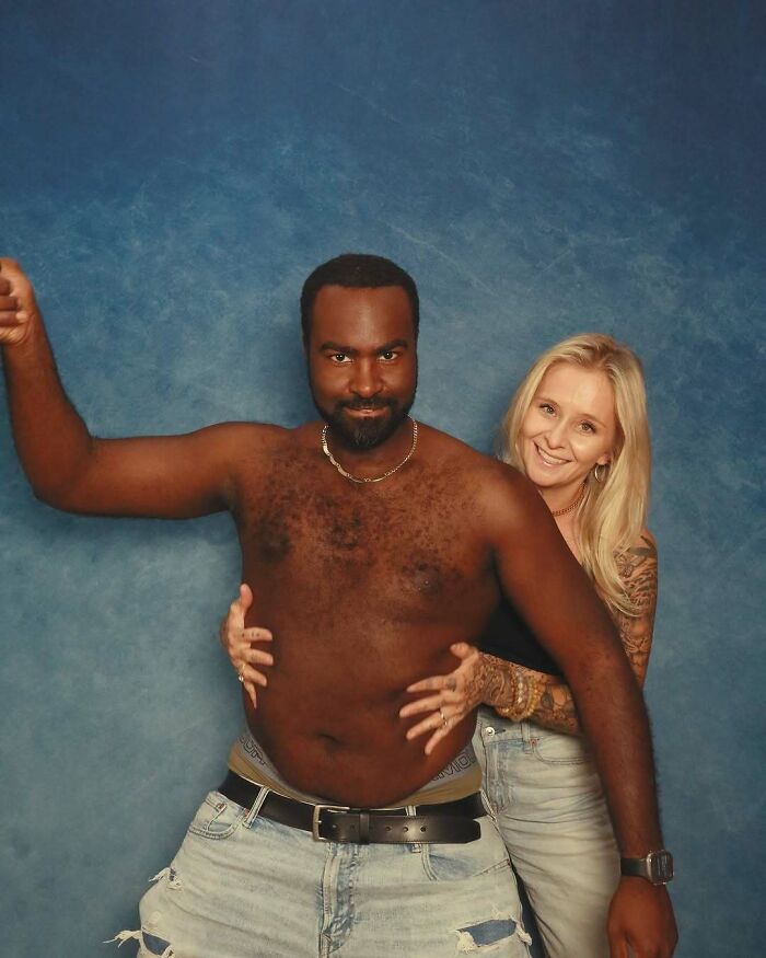 A shirtless man and a smiling woman hugging behind him in an awkward studio pics setting with a blue backdrop.