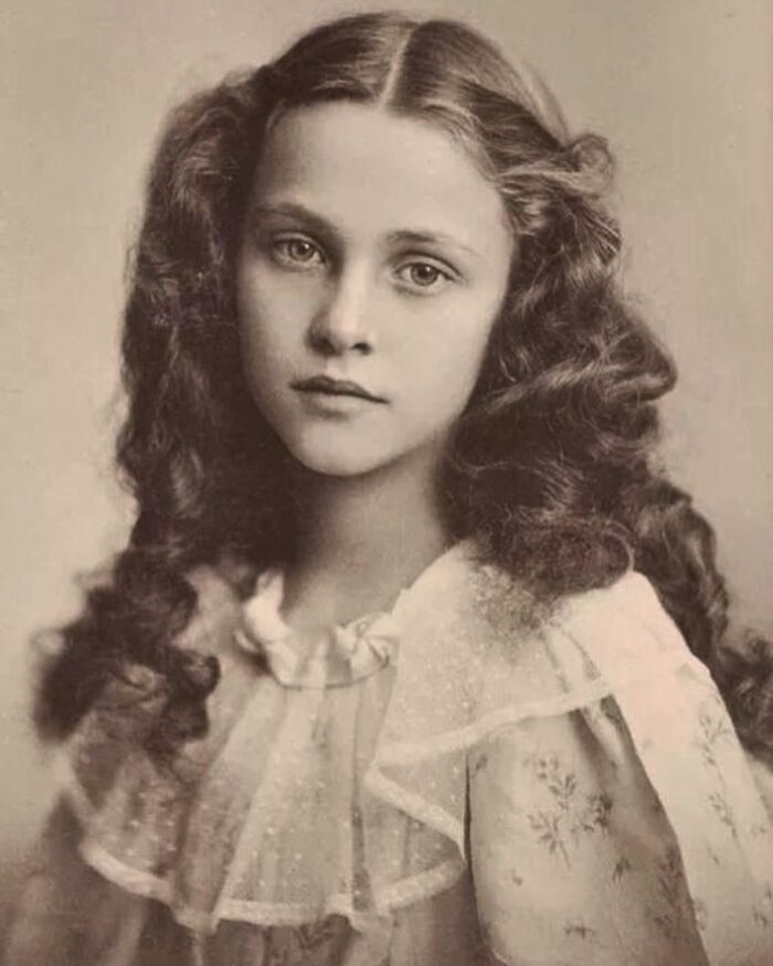 Young Victorian girl with long curly hair wearing a delicate ruffled dress in a weird Victorian era photo.