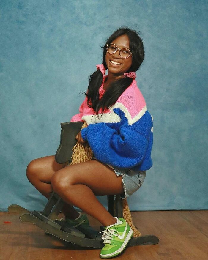 Young woman in colorful sweater and sneakers posing awkwardly on a rocking horse in a studio photo.