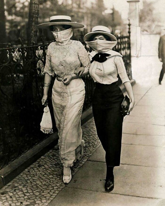 Two women wearing hats and face coverings walking arm in arm on a sidewalk in a weird Victorian era photo.