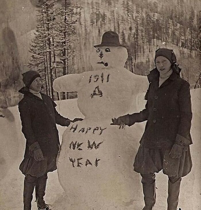 Two women standing beside a snowman with a hat, pipe, and text, in a weird Victorian era photo outdoors.