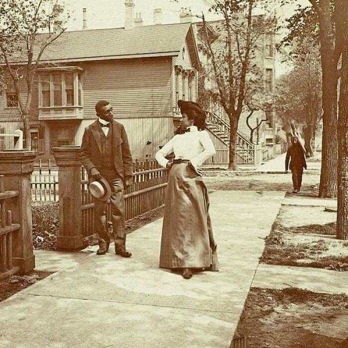 Victorian era photo of a man and woman in period clothing standing on a sidewalk near wooden fences and houses.