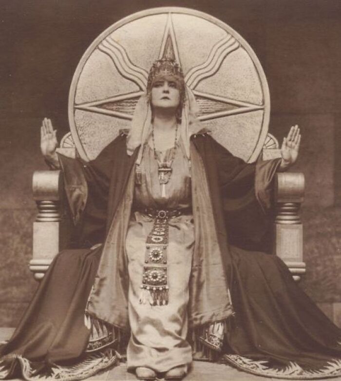 Victorian era woman in elaborate costume seated on ornate throne with raised hands in a weird Victorian era photo.