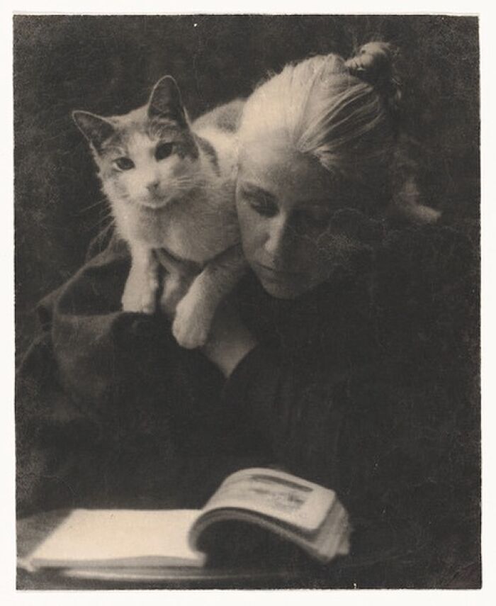 Victorian era photo of a woman with a cat on her shoulder, reading a book in a dimly lit setting.
