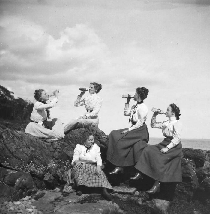 Four Victorian women in period clothing sitting on rocks, drinking from bottles in a weird Victorian era photo.