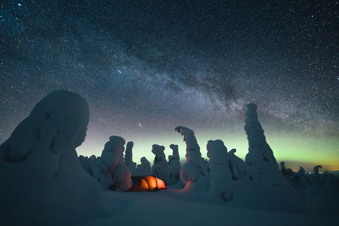 Snow-covered trees under a starry sky with the northern lights visible, and a glowing tent in a remote winter forest.