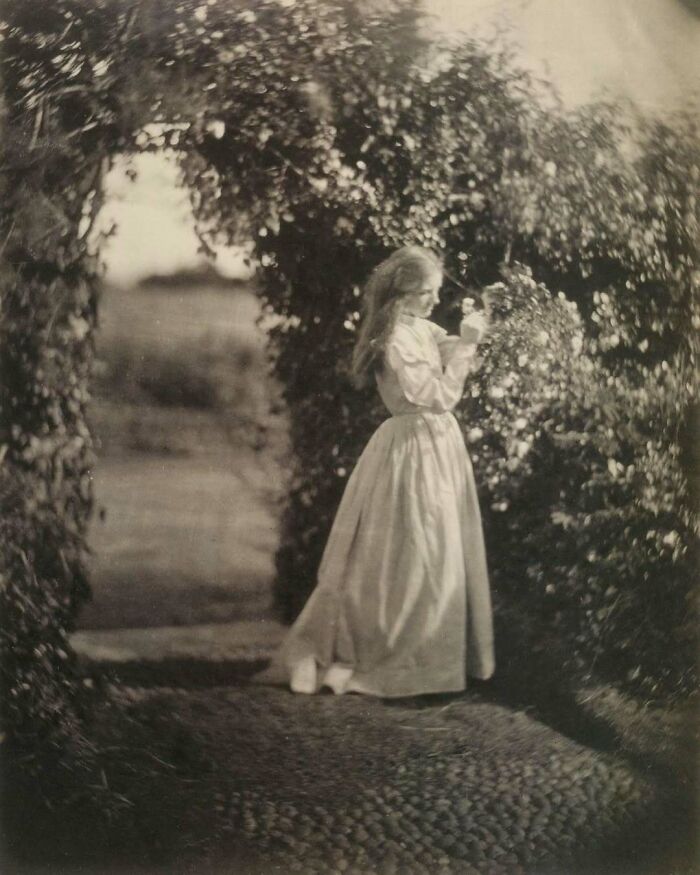 Victorian era photo of a young woman in a long dress standing under a leafy archway, highlighting weird Victorian era visuals.
