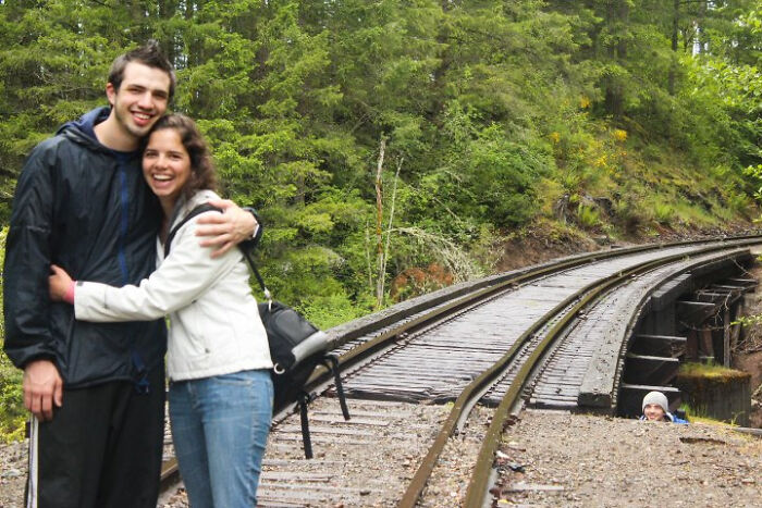 Couple hugging on railroad tracks while a person photobombs by peeking up from the ground nearby, epic photobombs humor.