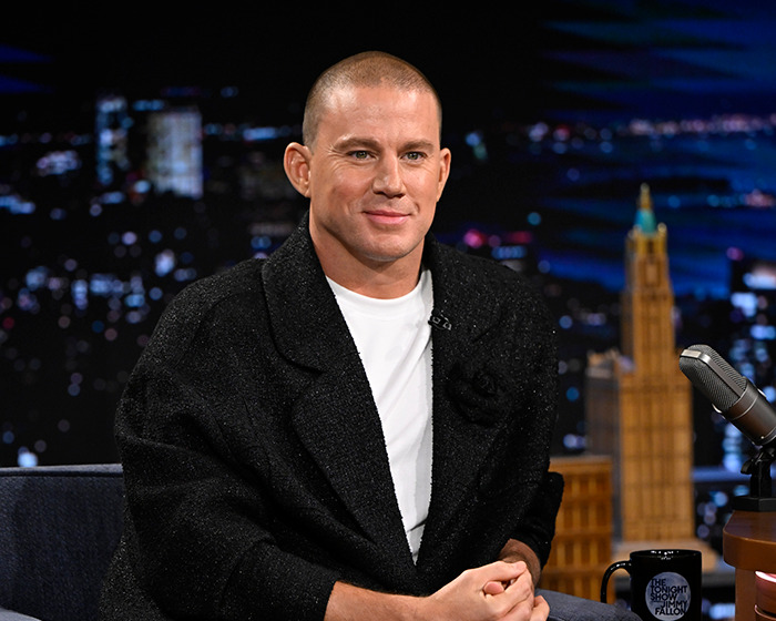 Actor sitting on a talk show set, wearing a black jacket, sharing stunning confessions about movies years after filming.