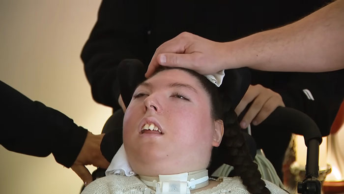 Young woman born without most of her brain lying in a wheelchair as family members care for her at home.
