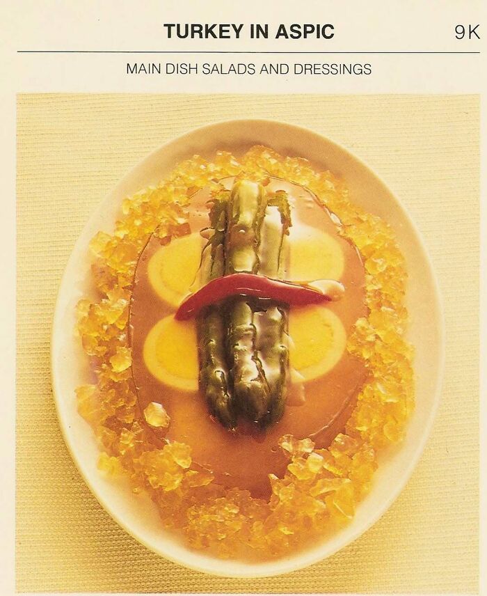 Vintage recipe of turkey in aspic surrounded by gelatin and garnished with pickles and a red pepper slice.