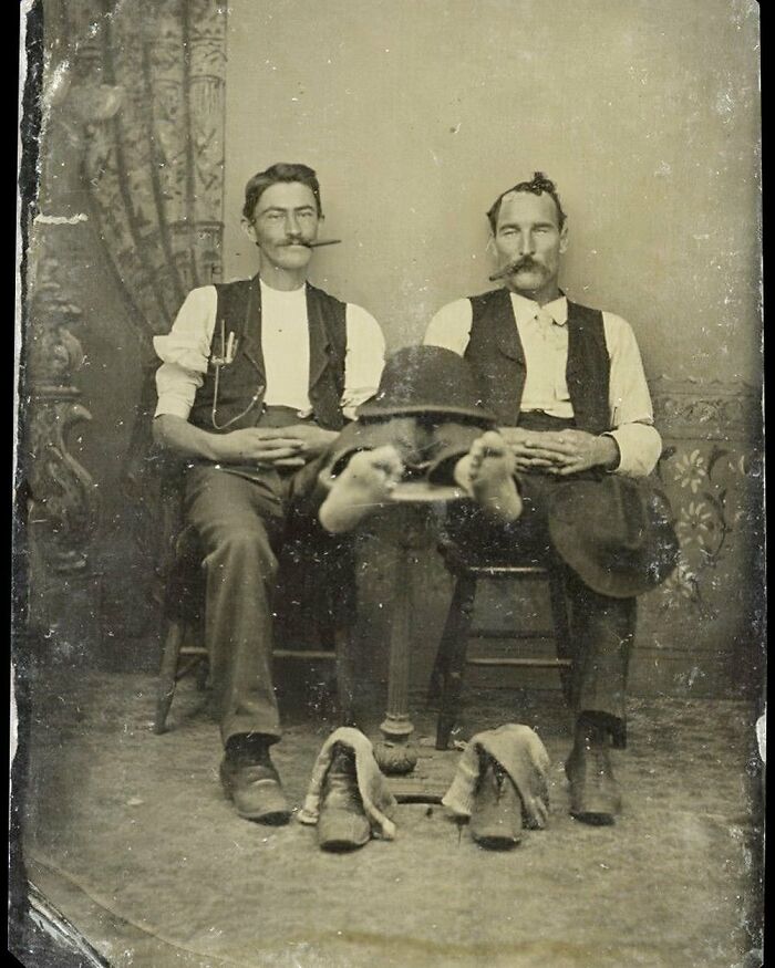 Two men with mustaches and cigars pose with a disembodied pair of feet, illustrating weird photos from the Victorian era.