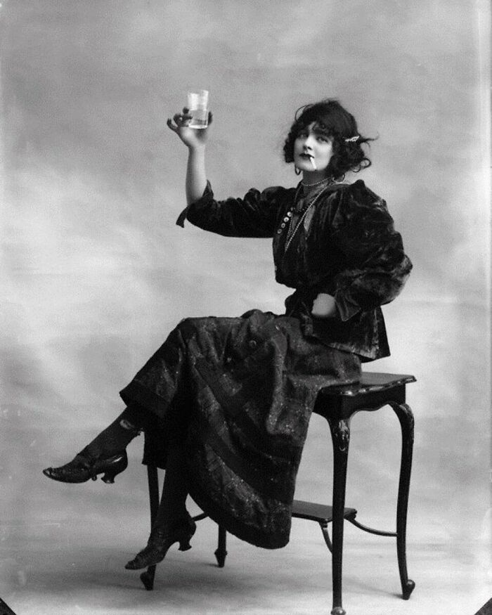 Woman in Victorian era clothing sitting on a table with a raised glass in a weird photo from the Victorian era.